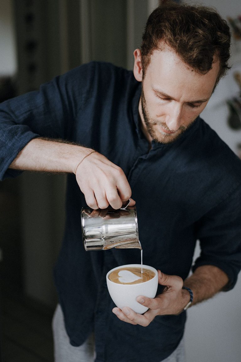 Mann in dunkelblauem Hemd mit Stehkragen macht Cappuccino mit Latte Art.