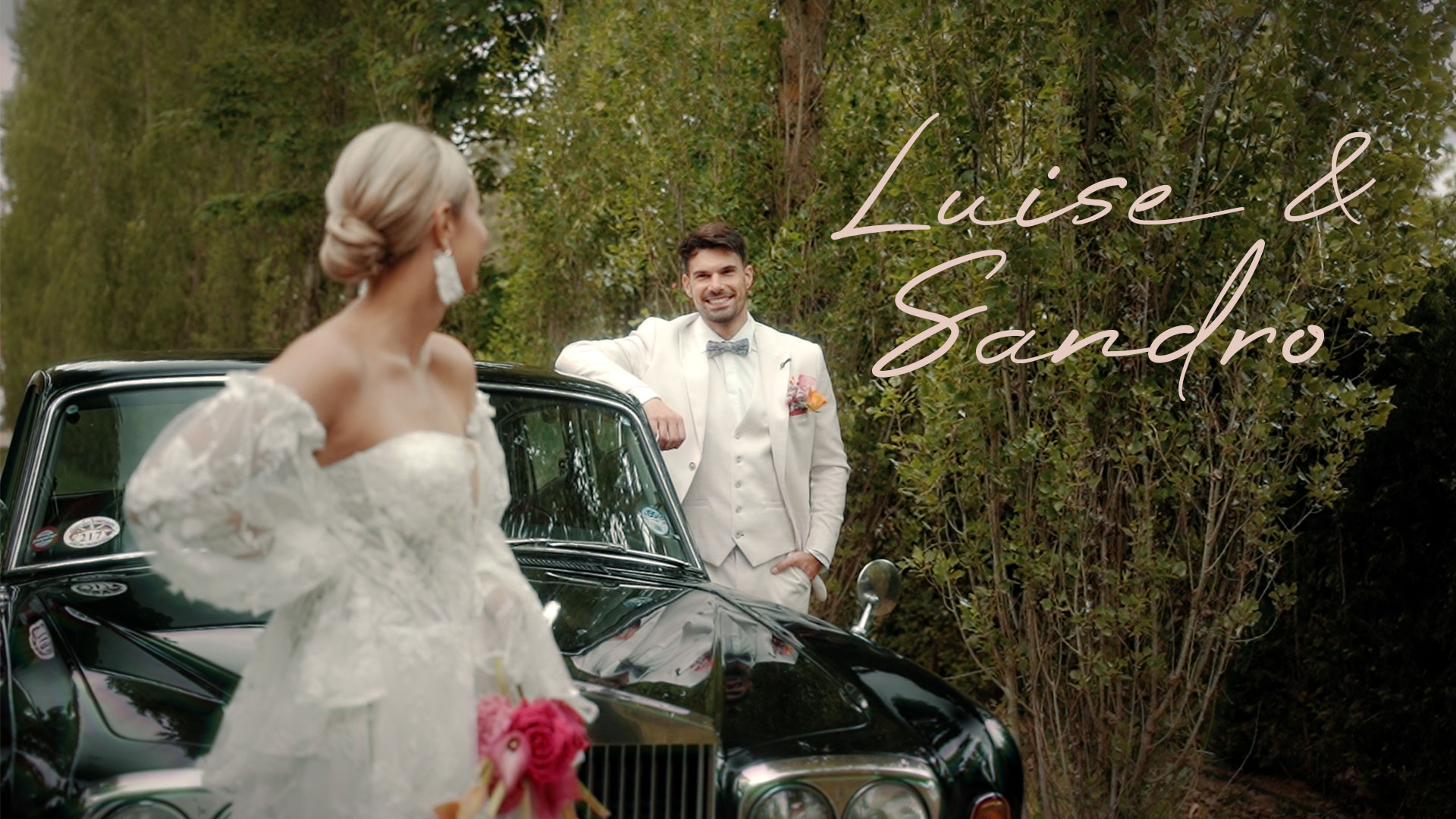 Hochzeitsfoto von Luise und Sandro: Die Braut steht im Vordergrund in einem eleganten Kleid mit Spitzenärmeln, der Bräutigam lehnt lächelnd an einem Oldtimer. Beide stehen auf einem Waldweg, rechts im Bild ihre Namen in eleganter rosa Schrift.