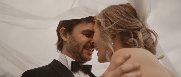 Nahaufnahme eines sich beinahe küssendem Brautpaars unter einem Schleier mit freudigem Lächeln - Hochzeitsvideos von Lukemotion